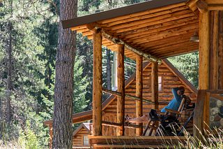A person relaxes in a chair on the shaded porch of a rustic log cabin, surrounded by tall pine trees.