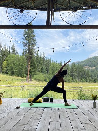 A person practices yoga on a covered wooden deck, with string lights overhead and a wide view of a meadow, pine forest, and mountains beyond.