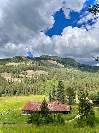 The south view from Minam Lodge, overlooking a grassy meadow and a large open shelter. Wooded hills are further south of the meadow. It is a sunny, cloudy day.