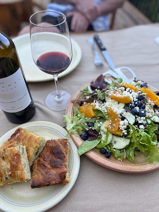 A colorful salad of blueberry, pear, orange slices, sunflower seeds, feta, on mixed greens next to a plate of focaccia and a bottle & glass of red wine.