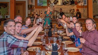 A group of smiling people at a large dining room table, holding their drinks up in a toast.