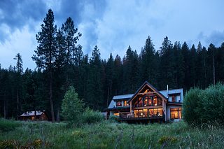 Minam River Lodge glows warmly at dusk from a grassy meadow, surrounded by tall evergreen forest beneath a blue, clouded sky.
