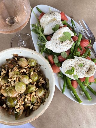 A salad platter with watermelon, green beans, burrata, arugula, and mint sits beside a bowl of roasted Brussels sprouts topped with toasted pine nuts.